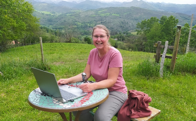 Vrouw zit buiten met laptop aan tafeltje in een grasveld tussen de bergen.