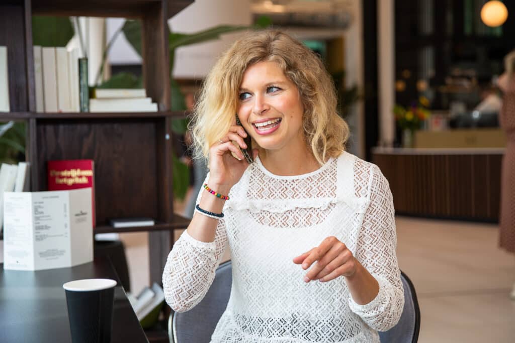 Blonde vrouw in wit shirt belt met haar smartphone en lacht.