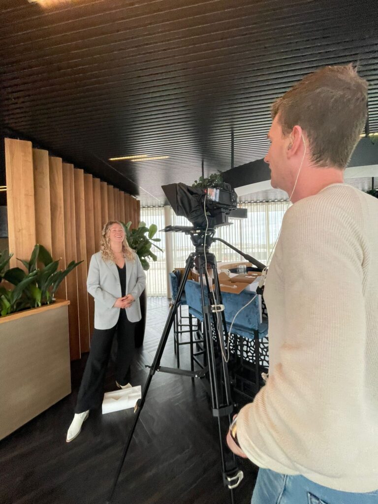 Renee staat te presenteren voor de camera, terwijl er een man achter de camera staat.