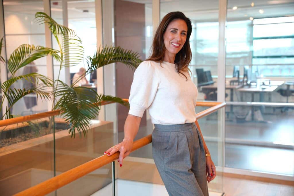 Tessa poseert lachend voor een houten reling. Op de achtergrond een kantoorruimte met bureaus en planten