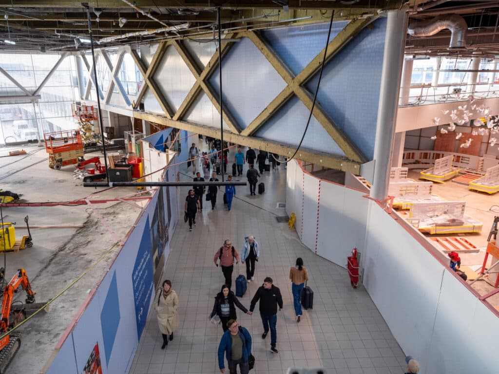 Een foto vanaf bovenaf gekomen. Reizigers lopen langs de bouwschotten in lounge 1. Achter de bouwschotten zijn bouwwerkzaamheden gaande.